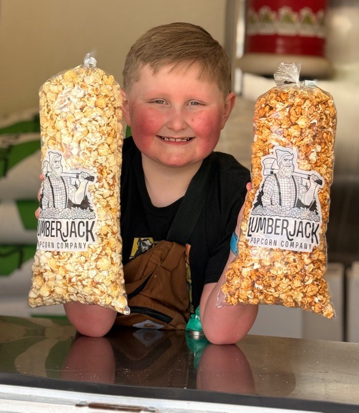 A smiling kid at the trailer window holding up bags of Original Kettle and Salted Caramel popcorn