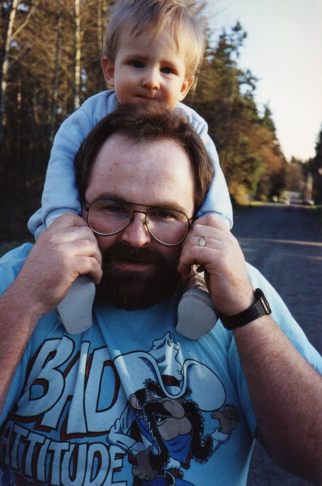 A young Greg carrying baby Nate on his shoulders down a tree-lined road